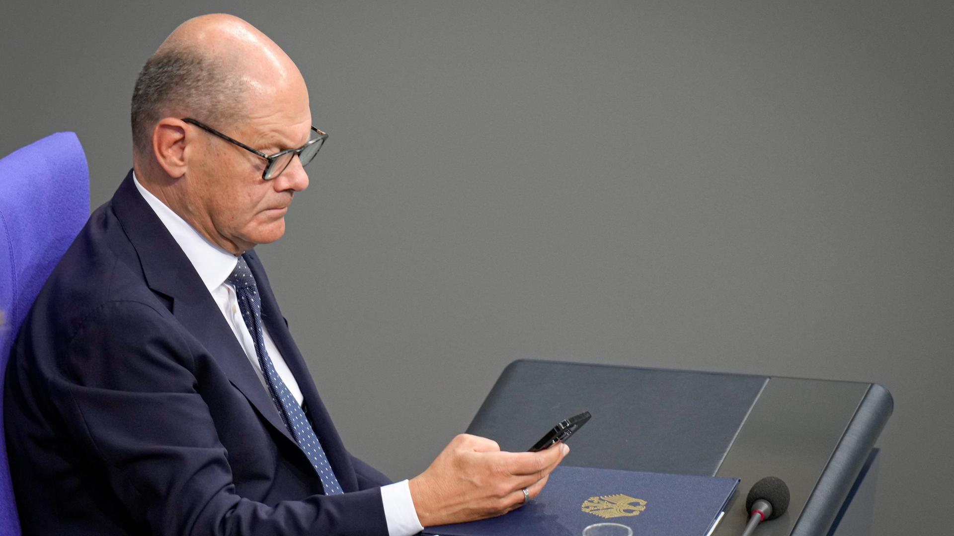 Bundeskanzler Olaf Scholz (SPD) hält im seitlichen Portrait auf der Regierungsbank sein Mobiltelefon in der Hand.