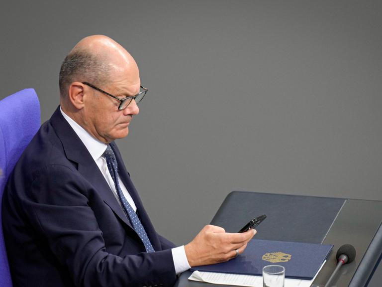 Bundeskanzler Olaf Scholz (SPD) hält im seitlichen Portrait auf der Regierungsbank sein Mobiltelefon in der Hand.