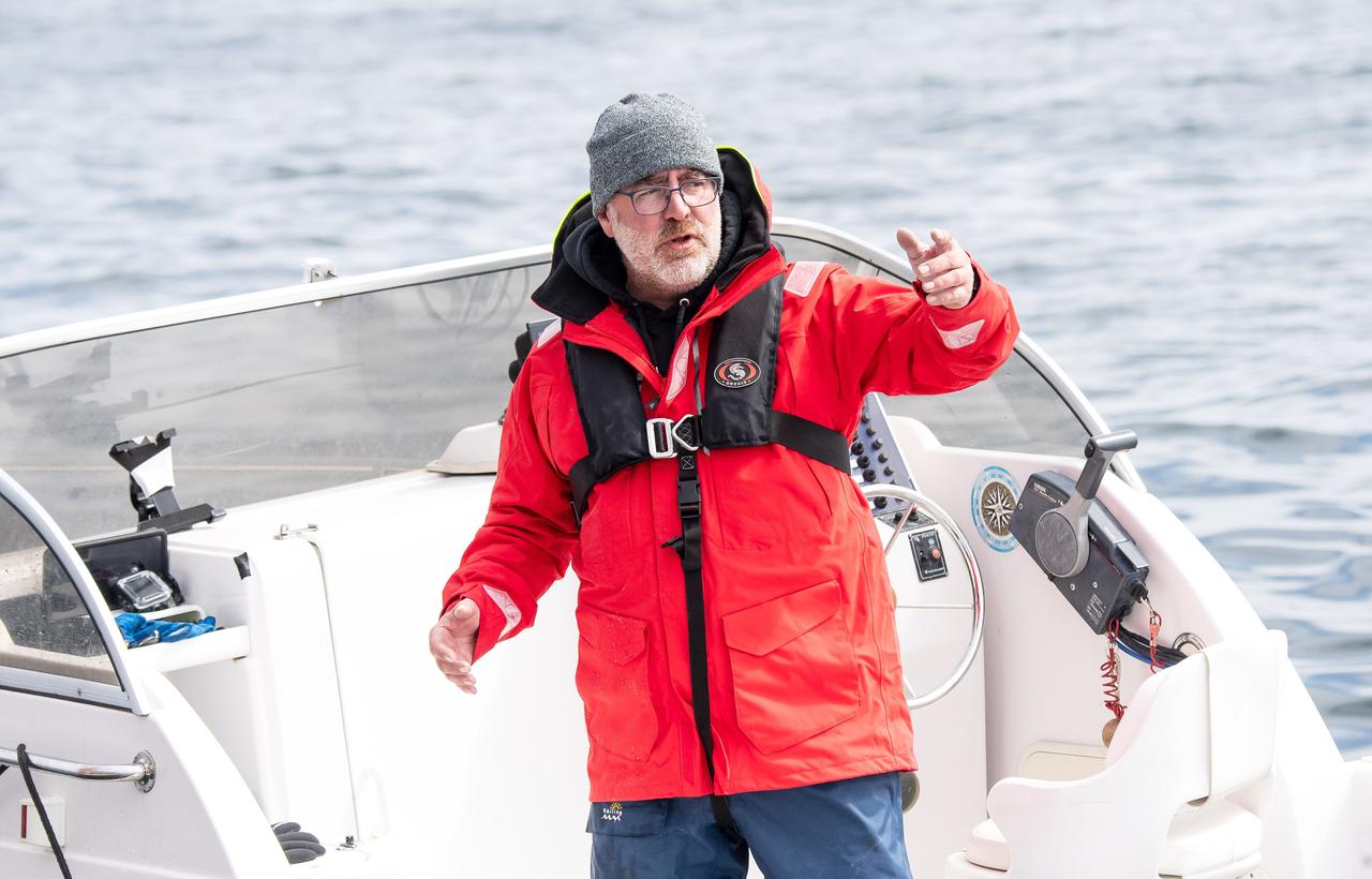 Ein Mamm in roter, wasserdichter Jacke, steht auf einem Boot und hält einen Arm auf Brusthöhe in der Luft. Er hat einen weißen Bart, trägt Brille und eine graue Wollmütze. 