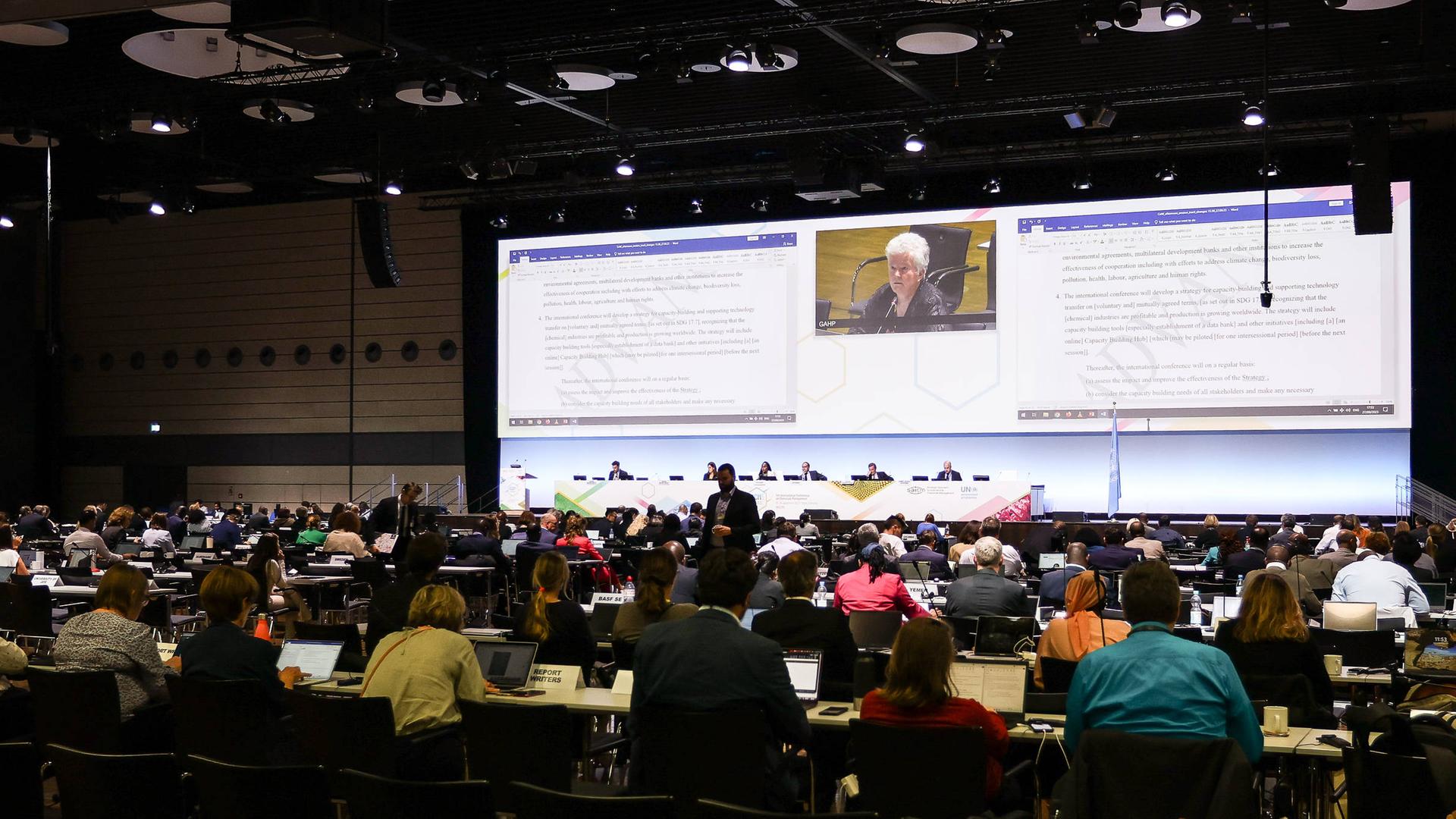 Blick in den Konferenzsaal mit Stuhlreihen, Bühne und großer Monitorwand.