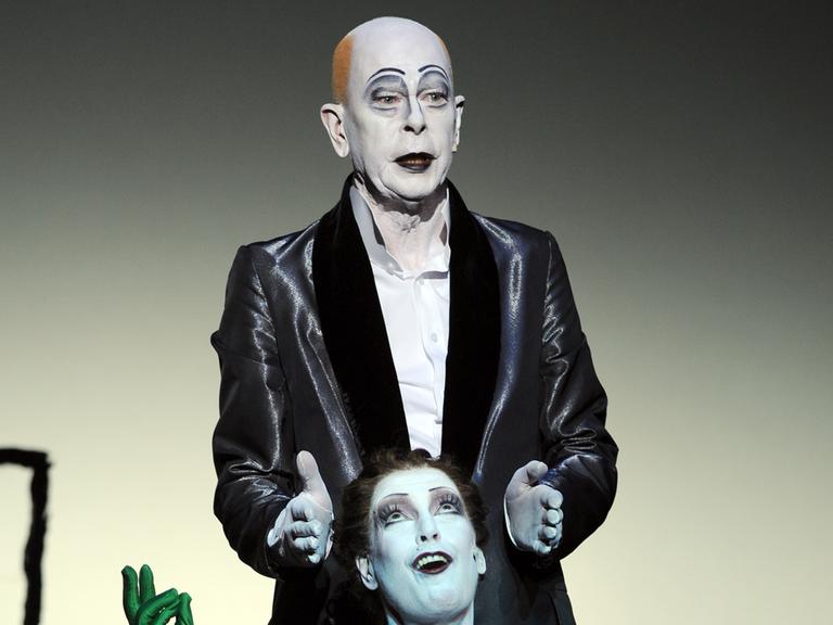 Der Schauspieler Alexander Lang auf der Theaterbühne mit einem grell geschminkten Gesicht.