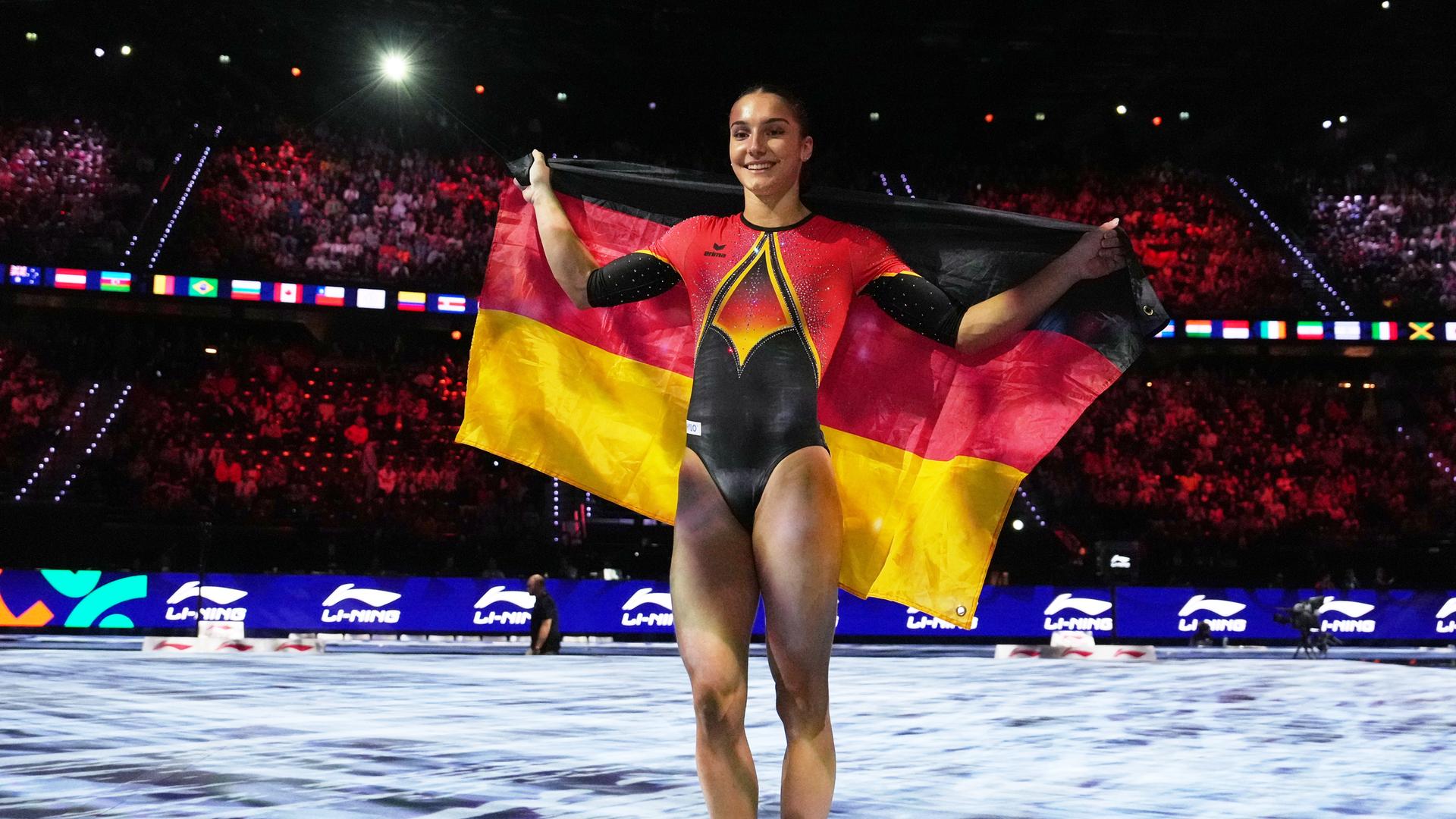 Zu sehen ist die deutsche Turnerin Helen Kevric. Sie steht auf der Wettkampffläche und hält die deutsche Flagge mit beiden Händen ausgebreitet hinter ihrem Rücken.