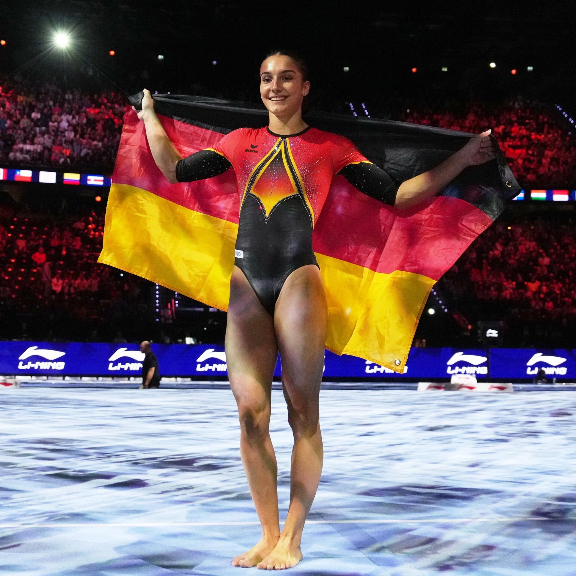 Zu sehen ist die deutsche Turnerin Helen Kevric. Sie steht auf der Wettkampffläche und hält die deutsche Flagge mit beiden Händen ausgebreitet hinter ihrem Rücken.