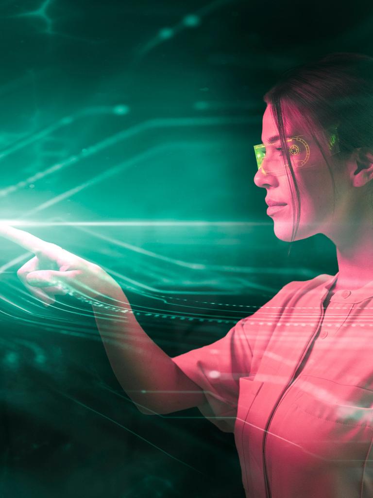 Eine junge Frau mit Laborbrille steht ihrem digitalem Spiegelbild gegenüber, in einer grünlich schimmernden Beleuchtung, es scheibt, als programmiert sie etwas (Fotomontage)