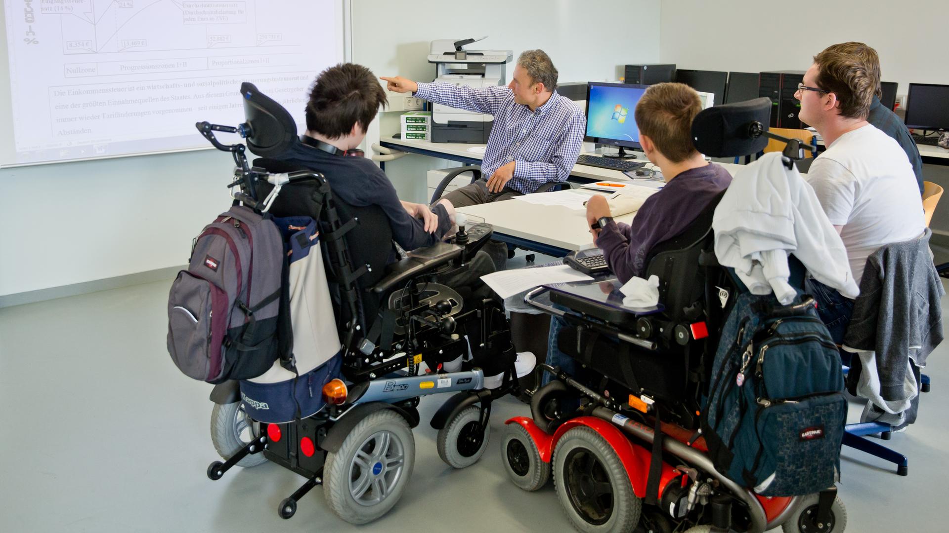 Zwei Schüler im Rollstuhl sitzen mit anderen Schülern an einem Tisch, ein Lehrer zeigt auf eine Projektion an der Wand. Zwei Schüler im Rollstuhl sitzen mit anderen Schülern an einem Tisch, ein Lehrer zeigt auf eine Projektion an der Wand.