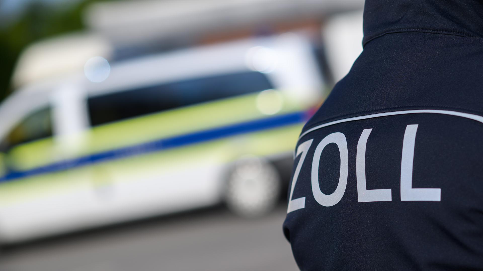 Ausschnitt einer Jacke mit dem Aufdruck "Zoll". Im Hintergrund ist verschwommen ein Polizeitransporter. 