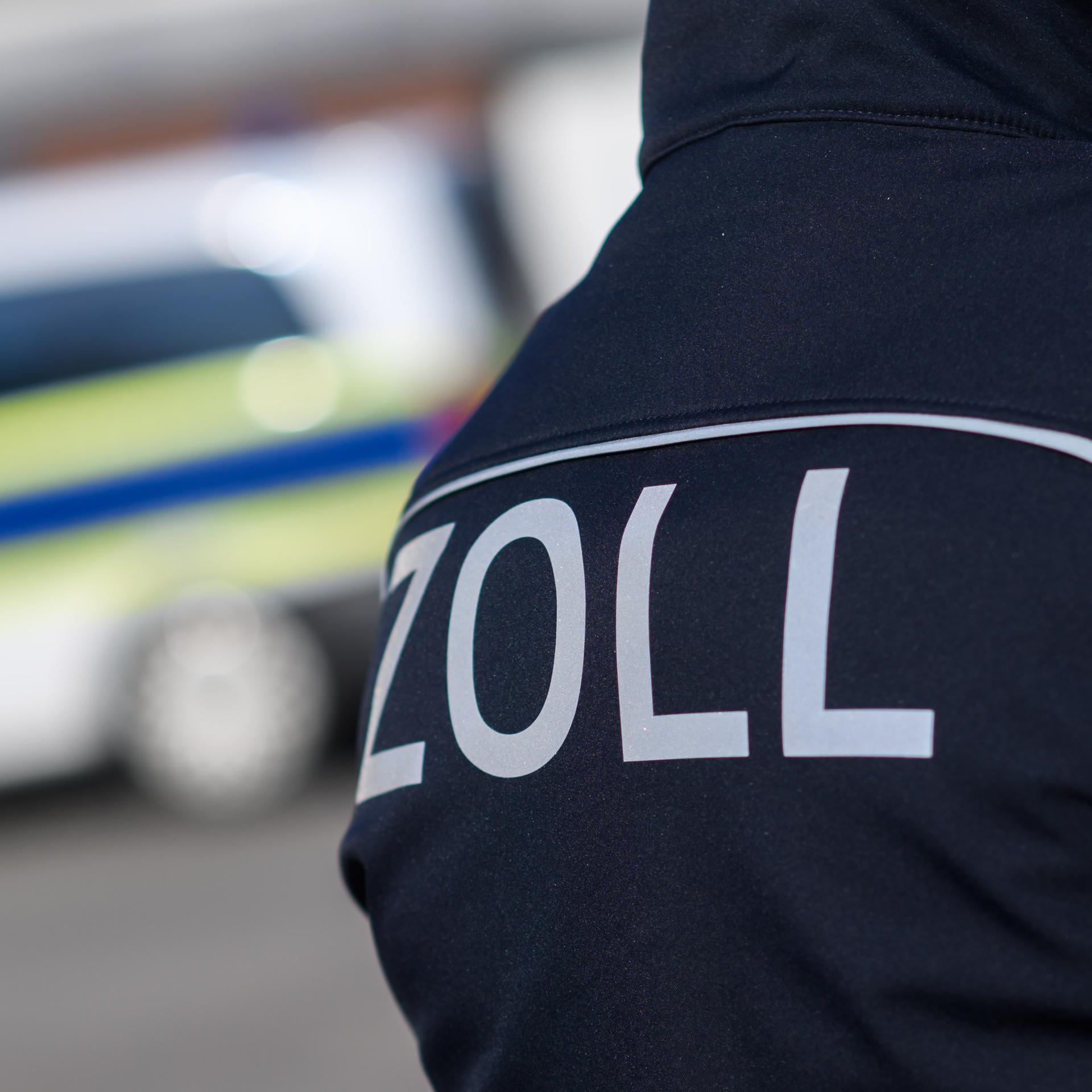 Ausschnitt einer Jacke mit dem Aufdruck "Zoll". Im Hintergrund ist verschwommen ein Polizeitransporter. 