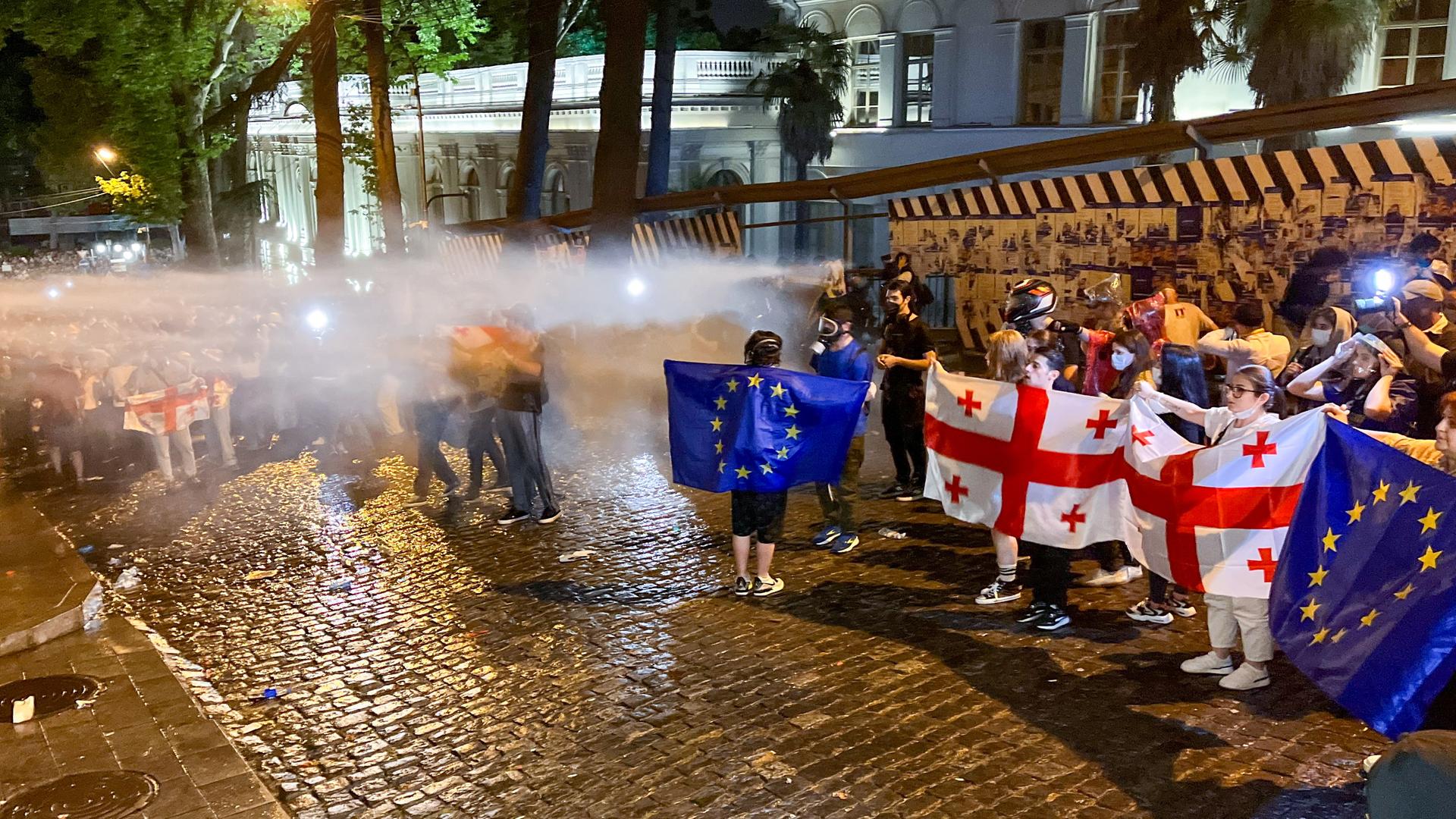 Demonstrierende in Tiflis halten georgische und EU-Flaggen hoch, während sie von der Polizei mit Wasser beschossen werden.