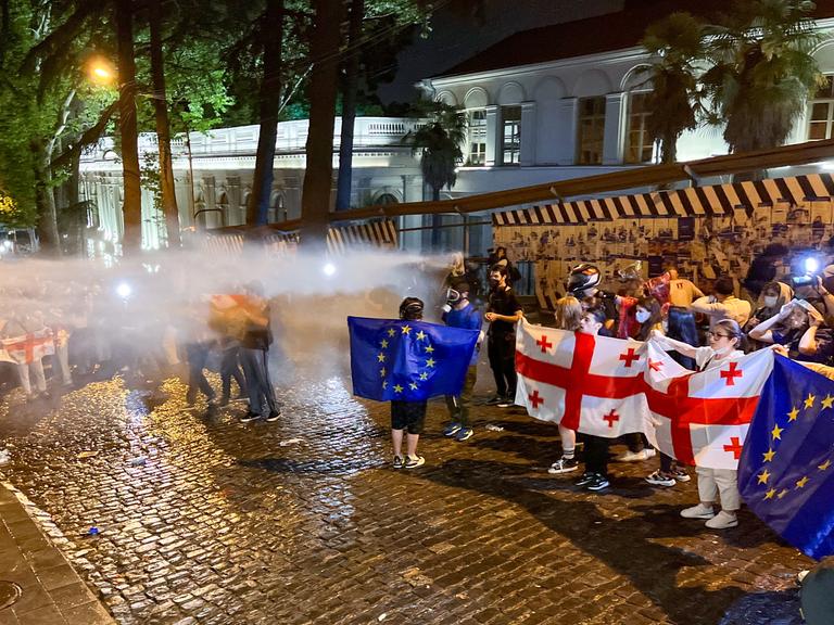 Demonstrierende in Tiflis halten georgische und EU-Flaggen hoch, während sie von der Polizei mit Wasser beschossen werden.