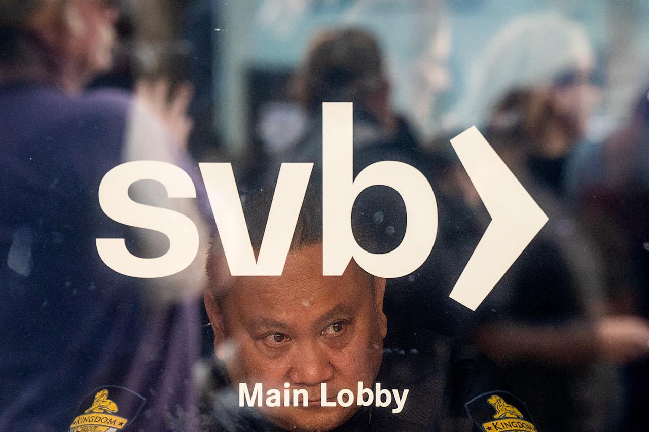 Ein Wachmann schaut durch die verglasten Türen der Filiale der Silicon Valley Bank in Santa Clara. Durch die Spiegelung der Scheibe ist zu sehen, dass sich draußen mehrere Menschen versammelt haben.