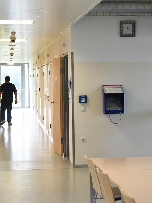Ein Pfleger bringt einen Maßnahmenhäftling auf sein Zimmer. Sie befinden sich in einem langen  in einem Gang mit dicht verschlossenen Türen, der Pfleger öffnet die Zimmertür des Häftlings.