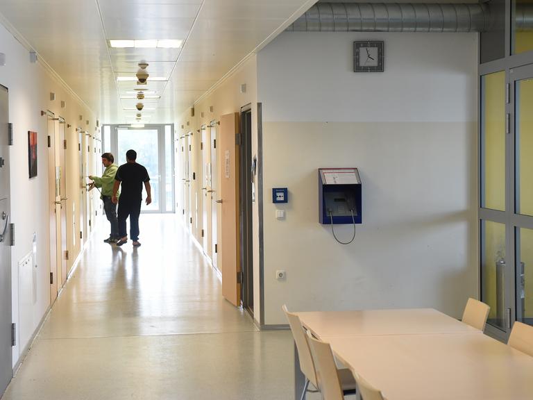 Ein Pfleger bringt einen Maßnahmenhäftling auf sein Zimmer. Sie befinden sich in einem langen  in einem Gang mit dicht verschlossenen Türen, der Pfleger öffnet die Zimmertür des Häftlings.