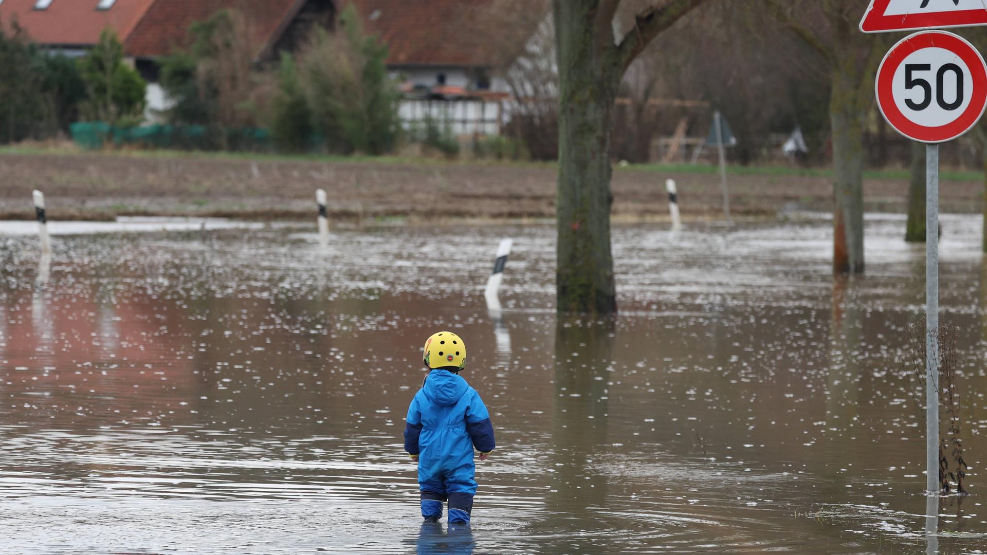 Lauenstadt bei Schulenburg (Region Hannover) ist im Dezember 2023 vom Hochwasser eingeschlossen. Ein Kleinkind in Gummistiefeln steht am Rand einer Straßenüberflutung, daneben ragt ein Verkehrsschild aus dem Wasser.