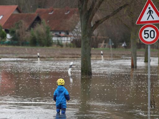 Lauenstadt bei Schulenburg (Region Hannover) ist im Dezember 2023 vom Hochwasser eingeschlossen. Ein Kleinkind in Gummistiefeln steht am Rand einer Straßenüberflutung, daneben ragt ein Verkehrsschild aus dem Wasser.