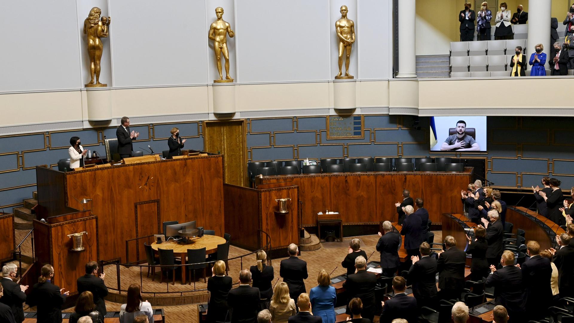 Blicks Parlament. Unten stehen im Halbrund die Abgeordneten, erhöht stehen Regierungsmitglieder. Auf einem großen Monitor ist Selenskyj zu sehen.