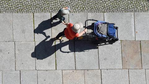 Mit Hut und Mütze als Sonnenschutz gehen eine Frau mit Kinderwagen und ein Kind einen Gehweg entlang.
