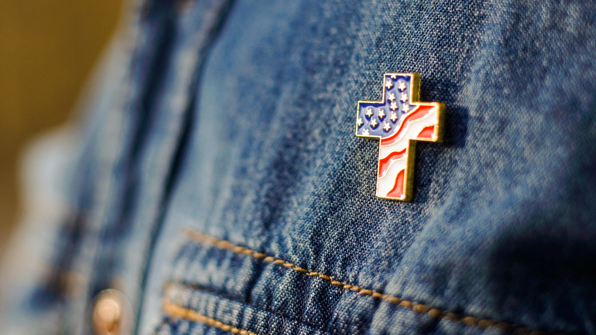 Eine Person mit Jeansjacke trägt einen Anstecker in Form eines Kreuzes, der mit den Stars and Stripes der amerikanischen Flagge gefüllt ist