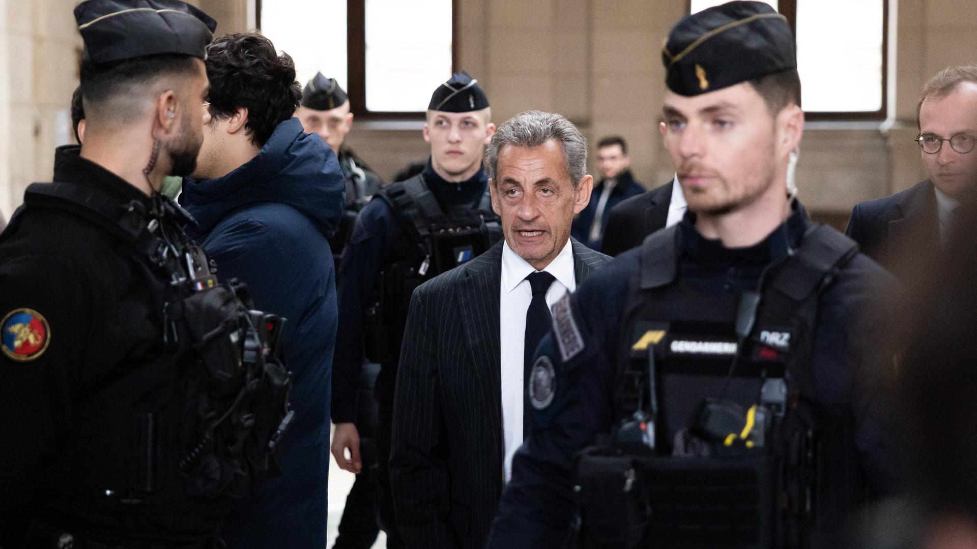 Frankreichs Ex-Präsident Nicolas Sarkozy im Februar 2024 vor Gericht in Paris. Auf dem Foto sind auch mehrere Polizeibeamte abgebildet.