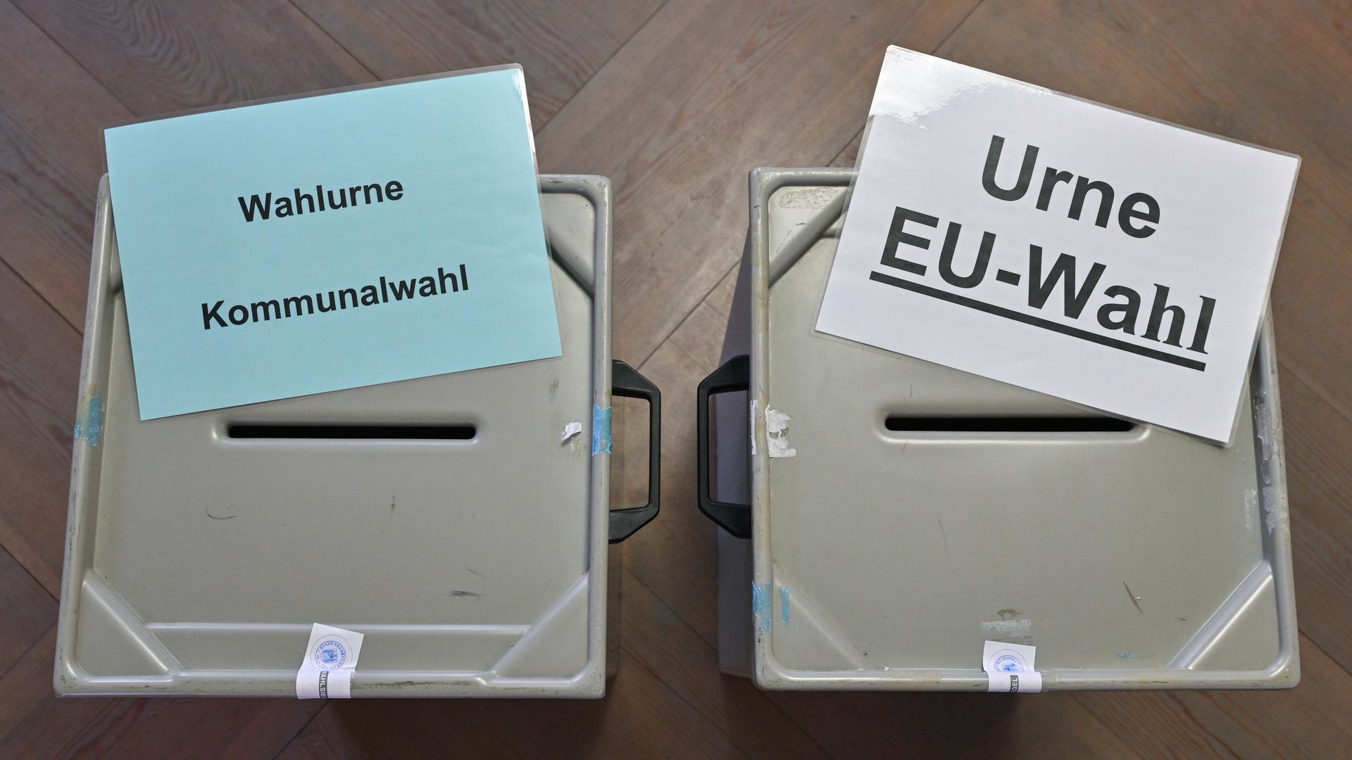 Zettel mit der Aufschrift "Wahlurne Kommunalwahl" (links) und "Urne EU-Wahl" (rechts) liegen auf zwei Wahlurnen in einem Wahllokal. Es sind graue Plastikbehälter mit einem Schlitz in der Oberseite.