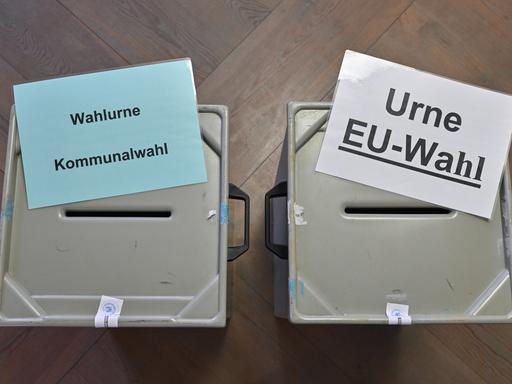 Zettel mit der Aufschrift "Wahlurne Kommunalwahl" (links) und "Urne EU-Wahl" (rechts) liegen auf zwei Wahlurnen in einem Wahllokal. Es sind graue Plastikbehälter mit einem Schlitz in der Oberseite.