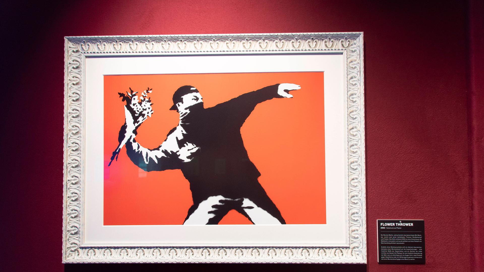 In einem Bilderrahmen ist ein Bild von Banksy zu sehen. Es zeigt in Schwarz einen Mann, der in der Haltung eines Steinewerfers nicht etwa Steine in der ausholenden Hand hält, sondern einen Blumenstrauß.