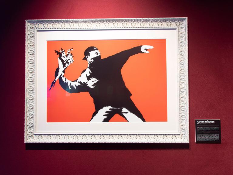 In einem Bilderrahmen ist ein Bild von Banksy zu sehen. Es zeigt in Schwarz einen Mann, der in der Haltung eines Steinewerfers nicht etwa Steine in der ausholenden Hand hält, sondern einen Blumenstrauß.