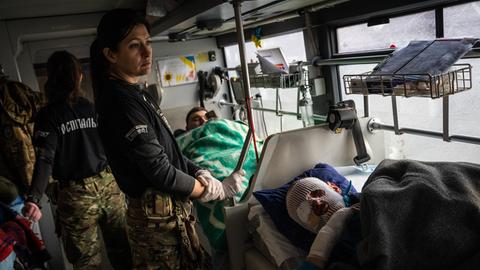 Das Bild zeigt einen verwundeten ukrainischen Soldaten mit Gesichtsverband, der von einer Sanitäterin in einem Großraumrettungswagen versorgt wird. 