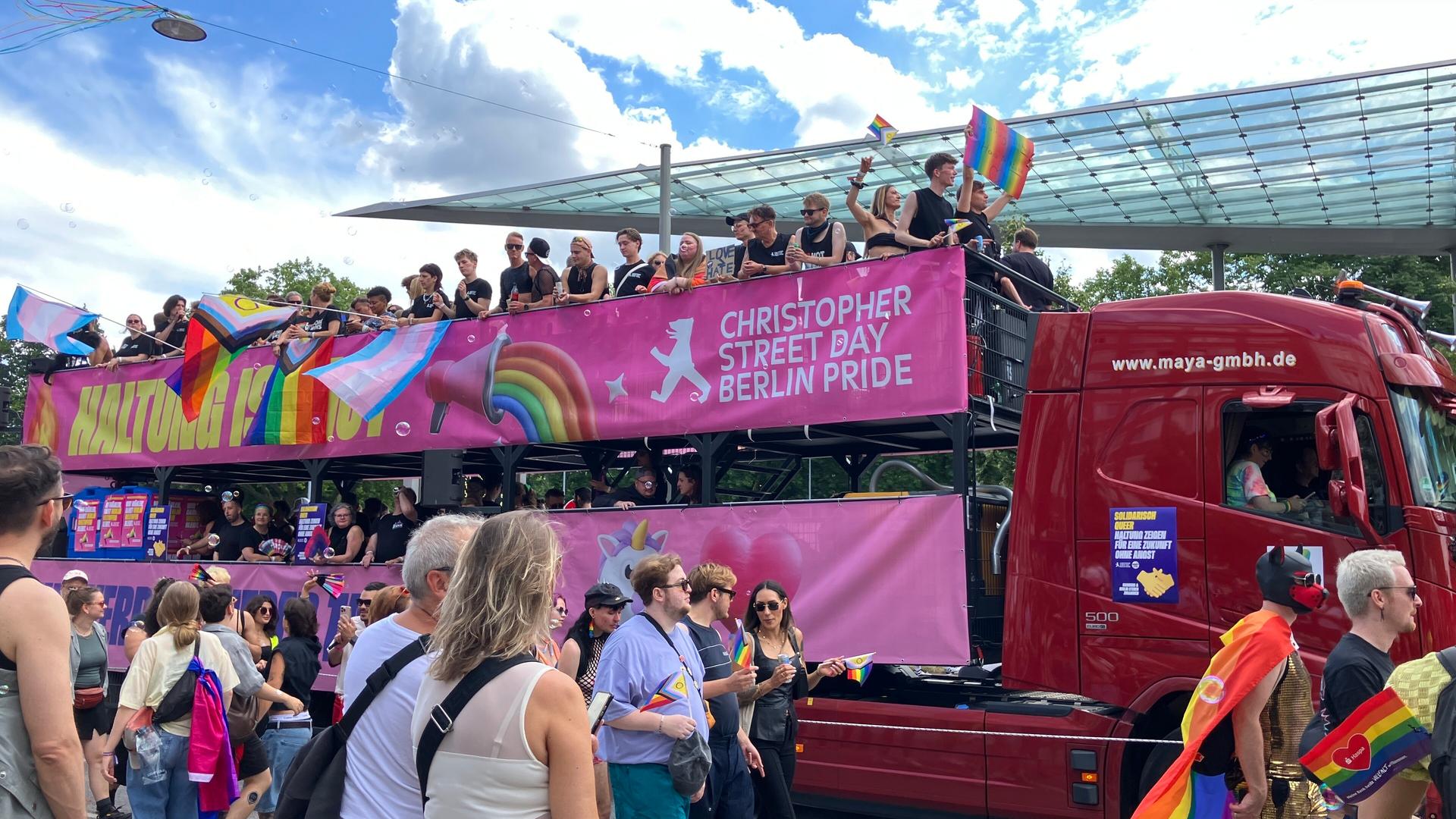 Auf dem Anhänger von einem Lastwagen stehen viele menschen und feiern. Auf einem Banner steht: Christopher Street Day Berlin Pride.