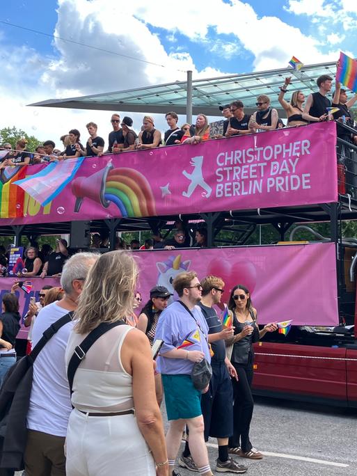 Auf dem Anhänger von einem Lastwagen stehen viele menschen und feiern. Auf einem Banner steht: Christopher Street Day Berlin Pride.