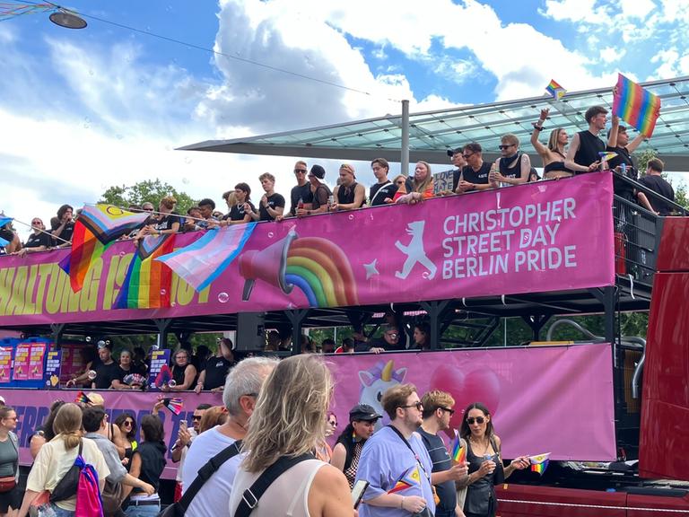 Auf dem Anhänger von einem Lastwagen stehen viele menschen und feiern. Auf einem Banner steht: Christopher Street Day Berlin Pride.