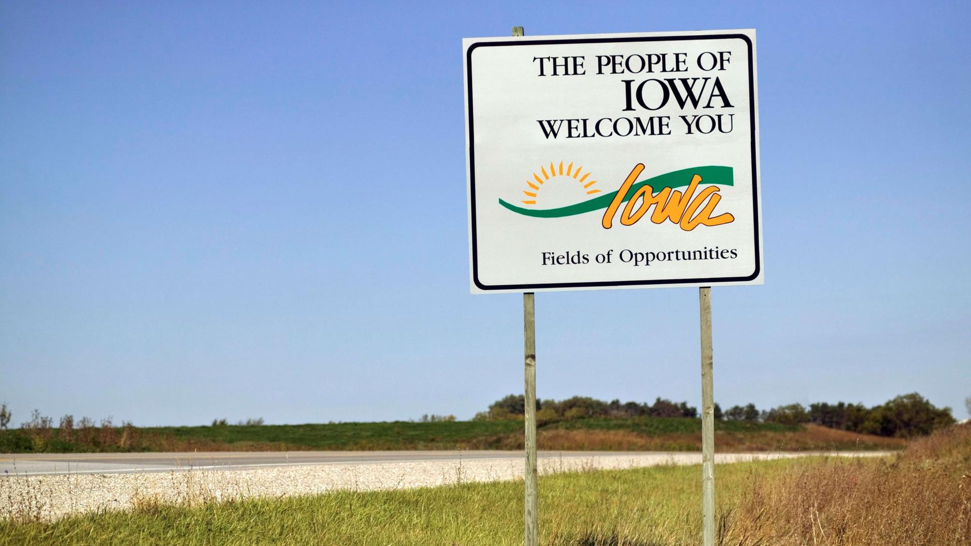 Ein Straßenschild "The People of Iowa welcome you" am Rand einer leeren Straße in der Landschaft von Iowa, USA.
