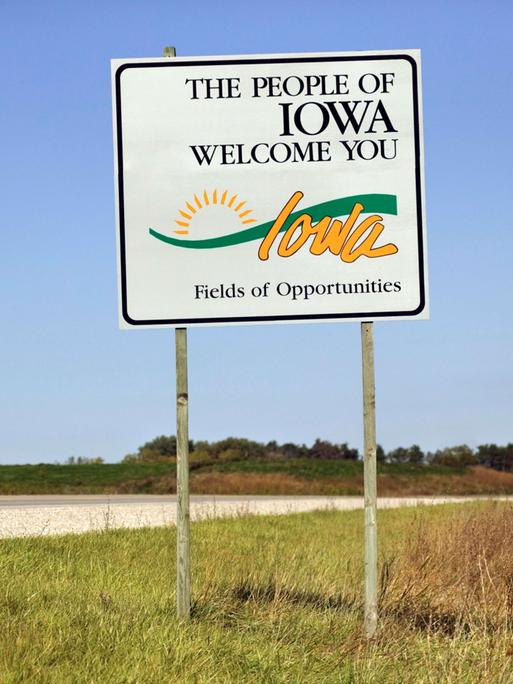 Ein Straßenschild "The People of Iowa welcome you" am Rand einer leeren Straße in der Landschaft von Iowa, USA.