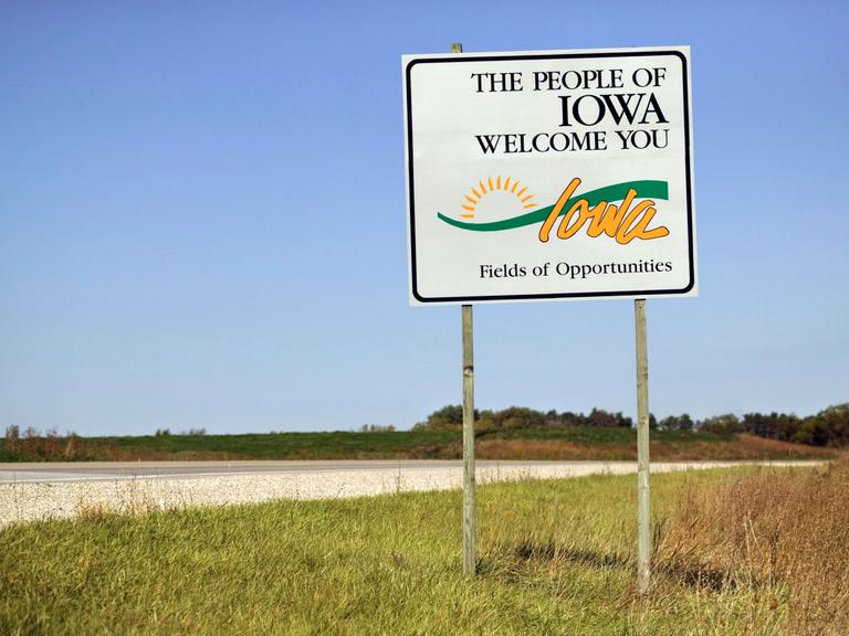 Ein Straßenschild "The People of Iowa welcome you" am Rand einer leeren Straße in der Landschaft von Iowa, USA.