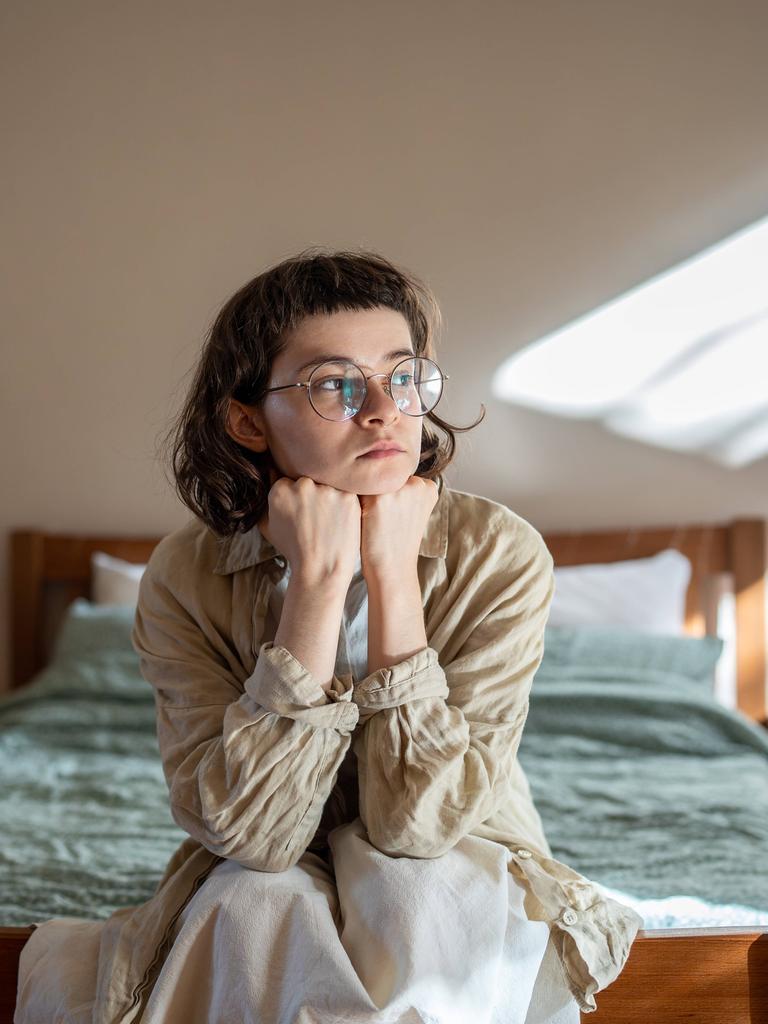 Depressive einsame junge Frau, die auf einem gemütlichen Bett sitzt und traurig auf das Fenster schaut und Angst hat. 