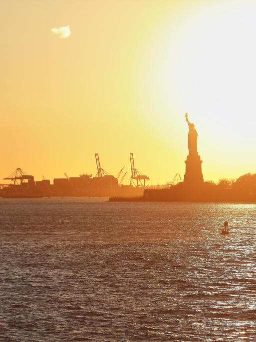 Der Hafen von New York City, USA mit seinen Kranen und Containerschifften, sowie der Freiheitsstatue bei Sonnenuntergang. 