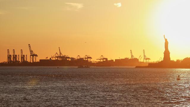 Der Hafen von New York City, USA mit seinen Kranen und Containerschifften, sowie der Freiheitsstatue bei Sonnenuntergang. 