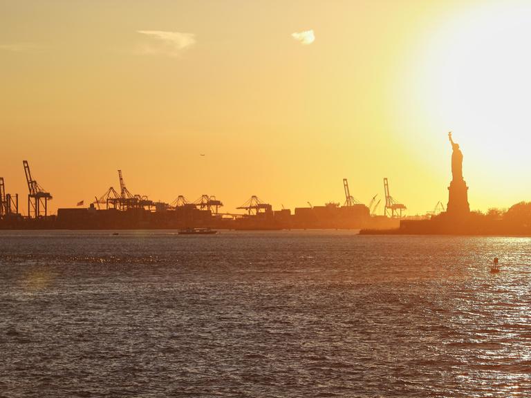 Der Hafen von New York City, USA mit seinen Kranen und Containerschifften, sowie der Freiheitsstatue bei Sonnenuntergang. 