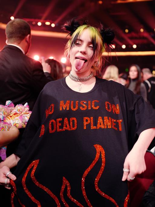 Billie Eilish streckt die Zunge heraus und zeigt ihr T-Shirt vor, auf dem in roten, pailettenbesetzten Buchstaben steht: "No Music On A Dead Planet".