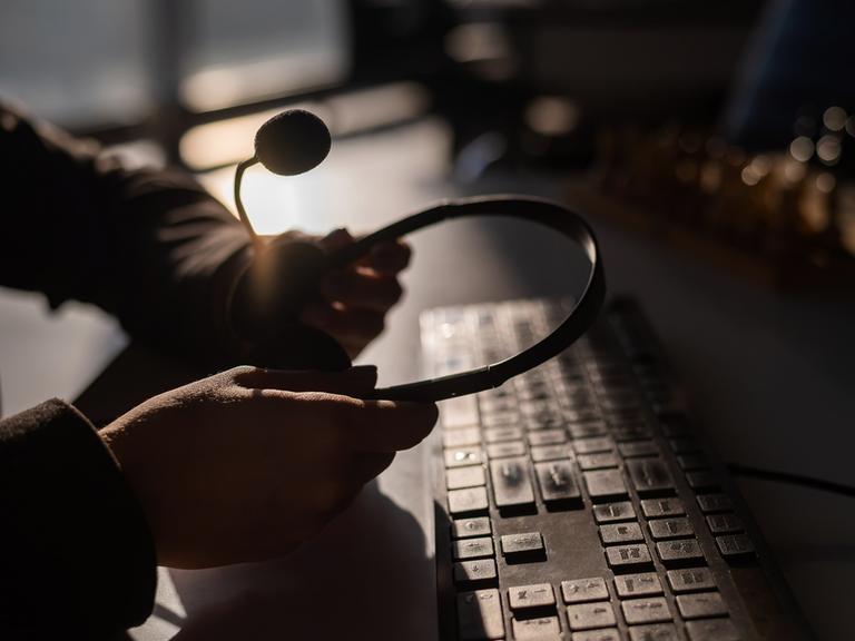 Dunkles Bild: Zwei Hände halten ein Headset über einer Tastatur.