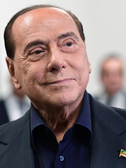 Porträt von Silvio Berlusconi, der an seinem blauen Anzug eine Anstecknadel in Form einer italienischen Flagge trägt.