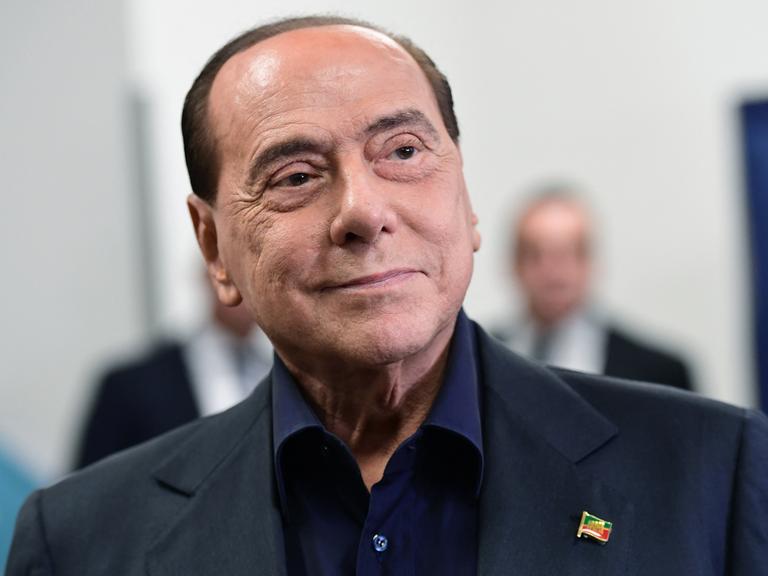 Porträt von Silvio Berlusconi, der an seinem blauen Anzug eine Anstecknadel in Form einer italienischen Flagge trägt.