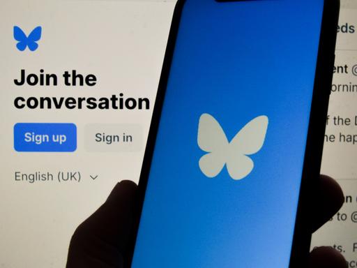 Ein Smartphone mit dem Logo der Plattform Bluesky. Es ist ein weißer Schmetterling auf blauem Hintergrund. 