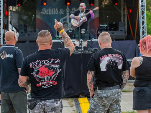 Vier Personen stehen bei einem Konzert vor der Bühne. Sie tragen Shirts mit Aufschriften wie "Rechtsrockland".