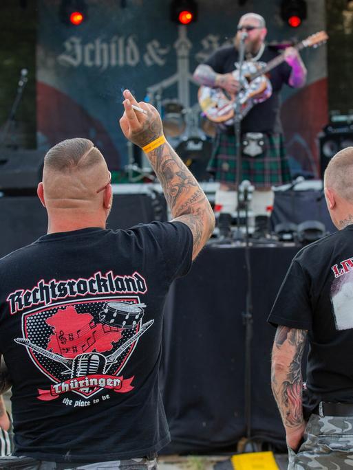 Vier Personen stehen bei einem Konzert vor der Bühne. Sie tragen Shirts mit Aufschriften wie "Rechtsrockland".
