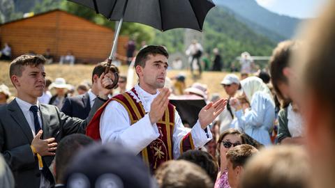 Ein Priester segnet in einer Menschenmenge eine Person. Eine andere Person hält einen Regenschirm als Sonnenschutz über den Priester.