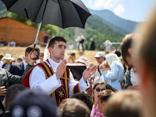 Ein Priester segnet in einer Menschenmenge eine Person. Eine andere Person hält einen Regenschirm als Sonnenschutz über den Priester.
