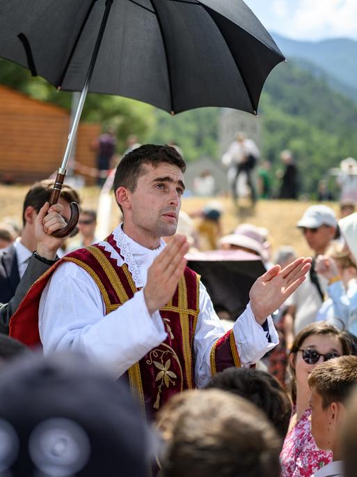 Ein Priester segnet in einer Menschenmenge eine Person. Eine andere Person hält einen Regenschirm als Sonnenschutz über den Priester.