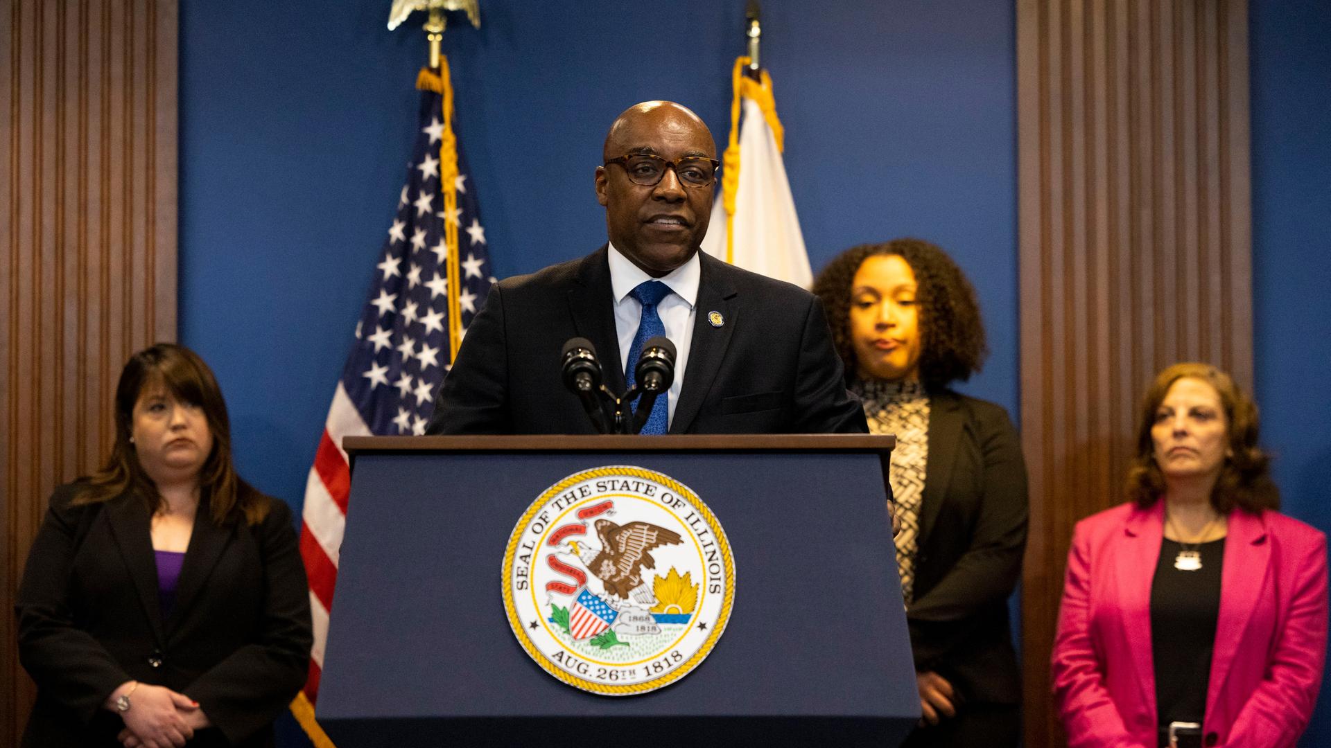 Der Generalstaatsanwalt von Illinois, Kwame Raoul, steht an einem Rednerpult mit dem Wappen des Bundesstaats Illinois. Vor einer dunkelblauen Wand stehen Flaggen der USA und des Bundesstaats. Hinter Raoul stehen mehrere Frauen.