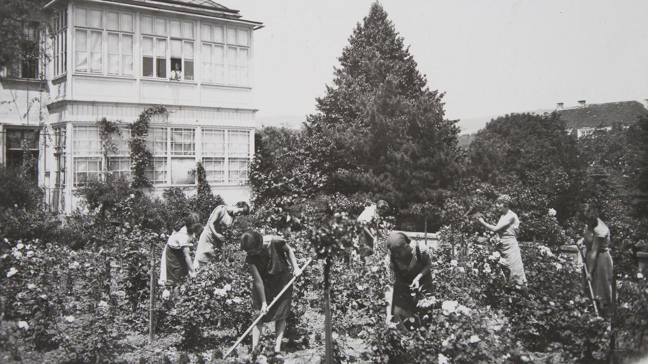 In einem Garten vor einem Haus kümmern sich junge Frauen um die Pflanzen.