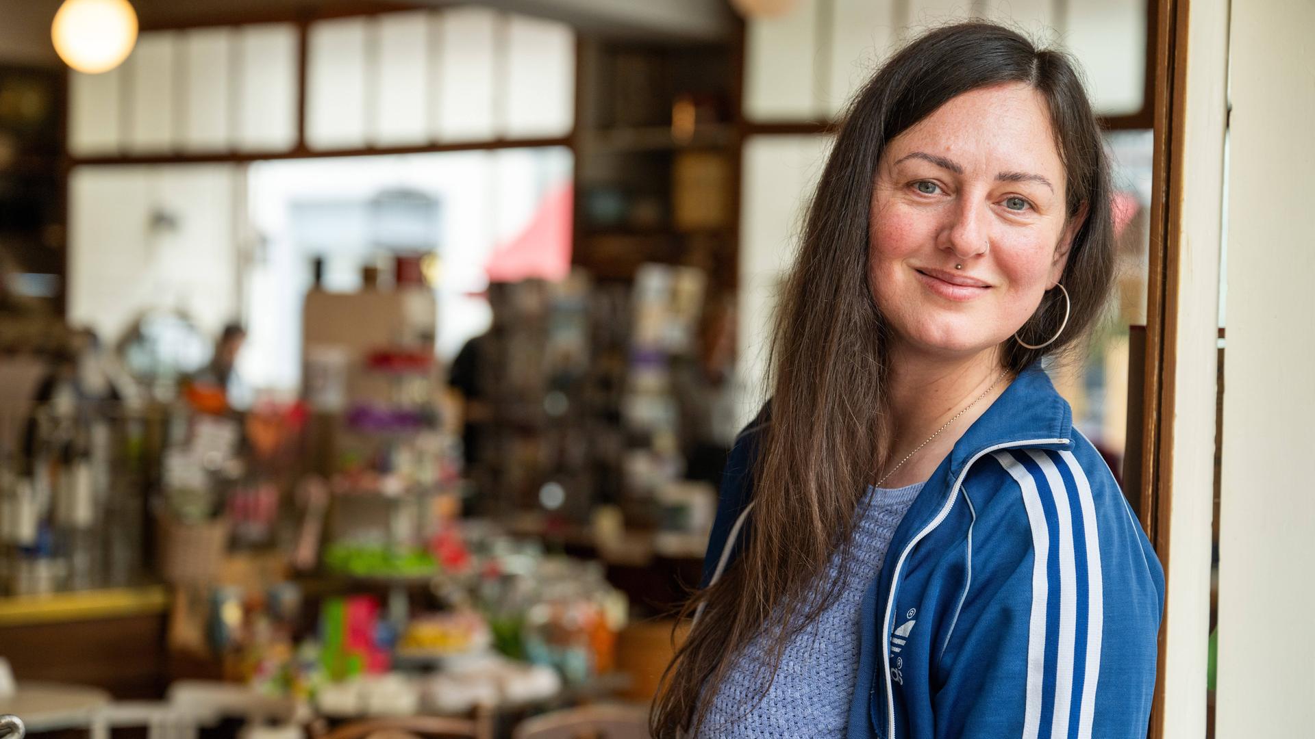 Nina Fuchs steht im Türrahmen eines Geschäfts und lächelt, sie trägt eine blaue Adidas-Jacke.